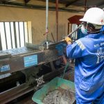Trabalhador usando macacão azul opera uma mesa vibratória para separar e processar minério em uma instalação industrial.