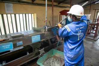 Trabalhador usando macacão azul opera uma mesa vibratória para separar e processar minério em uma instalação industrial.