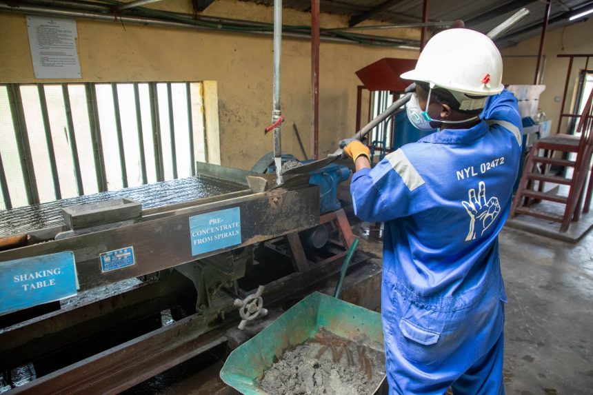 Trabalhador usando macacão azul opera uma mesa vibratória para separar e processar minério em uma instalação industrial.