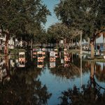Rua alagada com placas de interdição refletidas na água, entre árvores e calçadas.