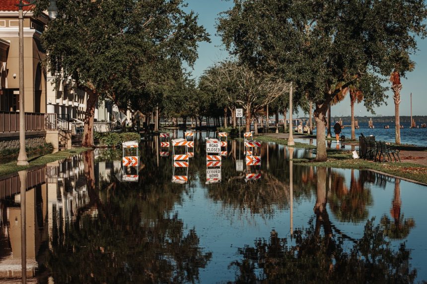 Rua alagada com placas de interdição refletidas na água, entre árvores e calçadas.