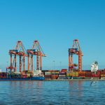 Porto de contêineres com guindastes e navios em uma área industrial.