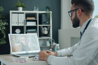 Médico de jaleco realiza uma consulta por videochamada com um paciente em um laptop, em um ambiente de atendimento moderno.