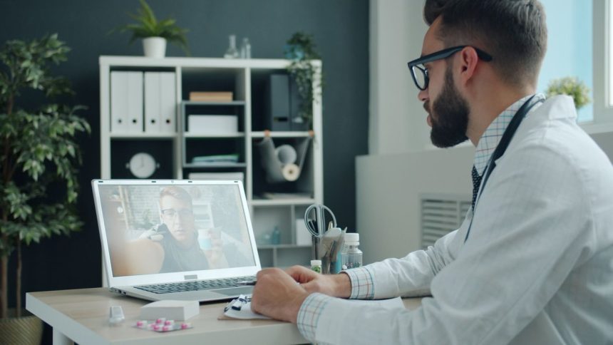 Médico de jaleco realiza uma consulta por videochamada com um paciente em um laptop, em um ambiente de atendimento moderno.