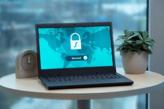 Laptop com ícone de cadeado na tela, sobre uma mesa ao lado de uma planta e um relógio.