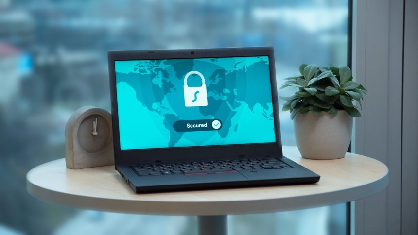 Laptop com ícone de cadeado na tela, sobre uma mesa ao lado de uma planta e um relógio.