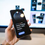 Pessoa segura um smartphone com um aplicativo de carteira digital aberto, enquanto um monitor aparece desfocado ao fundo.