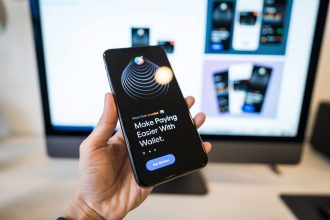 Pessoa segura um smartphone com um aplicativo de carteira digital aberto, enquanto um monitor aparece desfocado ao fundo.