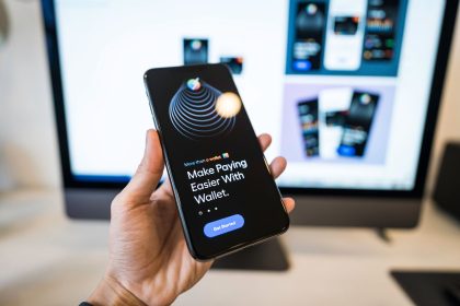 Pessoa segura um smartphone com um aplicativo de carteira digital aberto, enquanto um monitor aparece desfocado ao fundo.